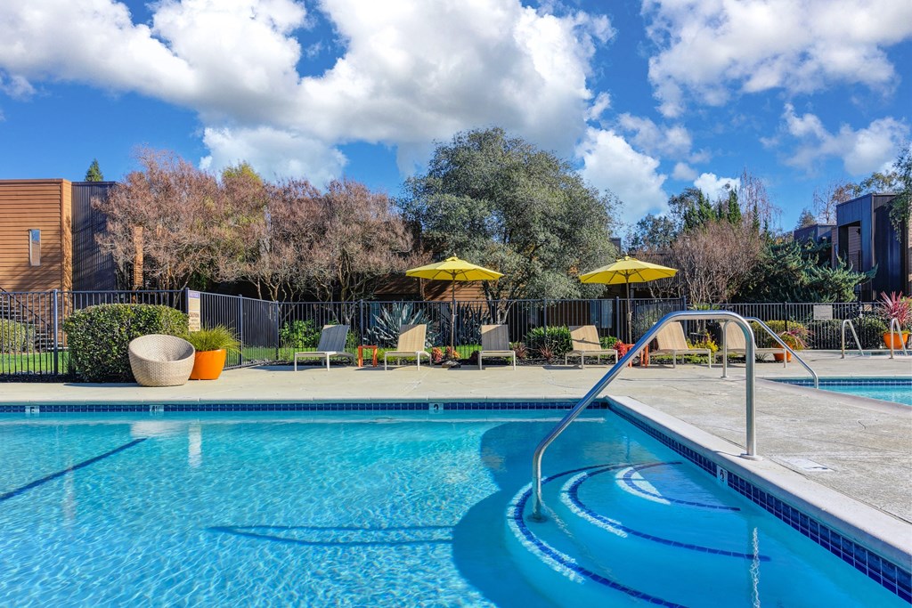 Swimming pool entrance. Three steps leading into the clear blue water. Bright yellow umbrellas and lounge chairs on the pool deck area.at Fountains of Fair Oaks, Fair Oaks, CA