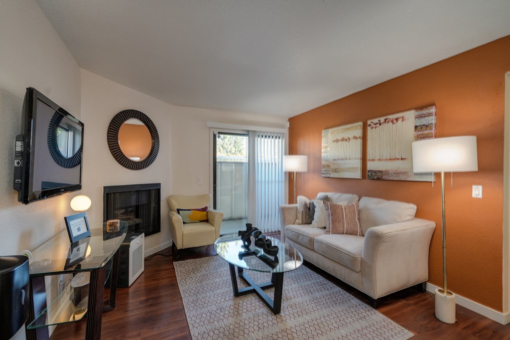 Model Home Living Room with Hardwood Inspired Floor, Rug, Glass Table, Round Wall Mounted Mirror above the gas fireplace, White/Gray Sofa, and Flat Screen Television