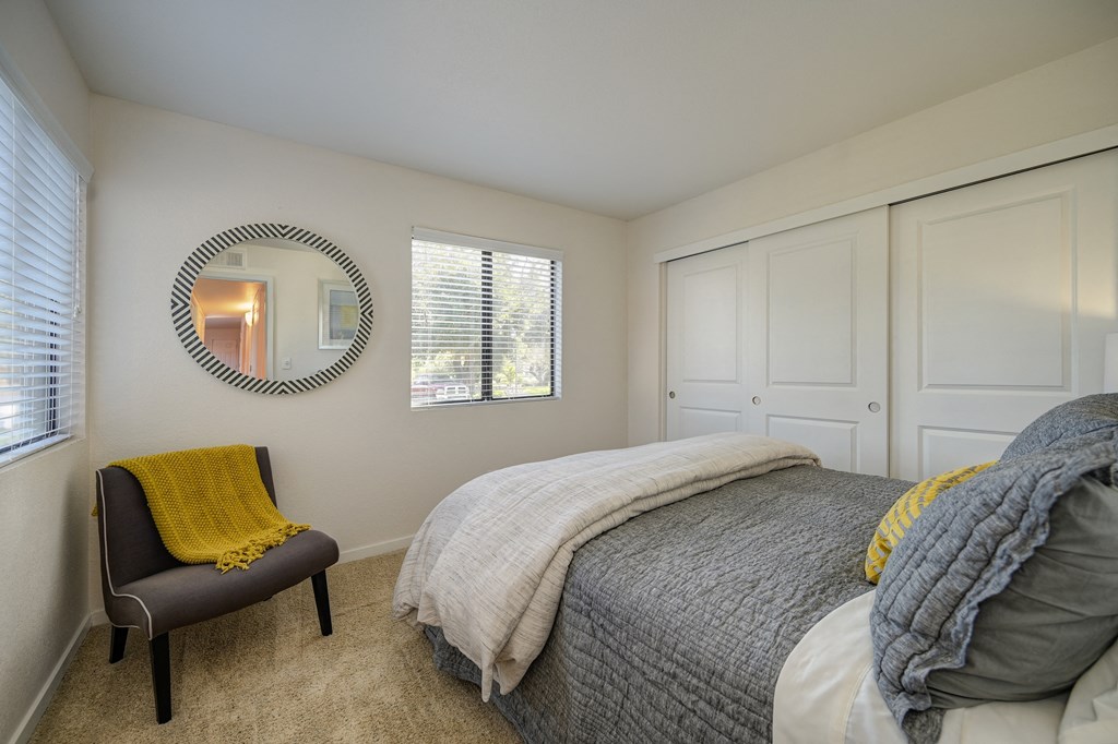 Master Bedroom with gray comforter. Room has Carpet, Gray Chair in corner, large closet with 3 doors and Window at Rocklin Manor  Apartments, California