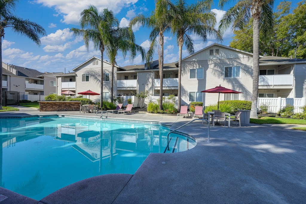 Community swimming pool with large palm trees in the background as well as one of the community apartment buildings. There are tables and chairs around the pool deck at The Legacy Apartments, Antelope