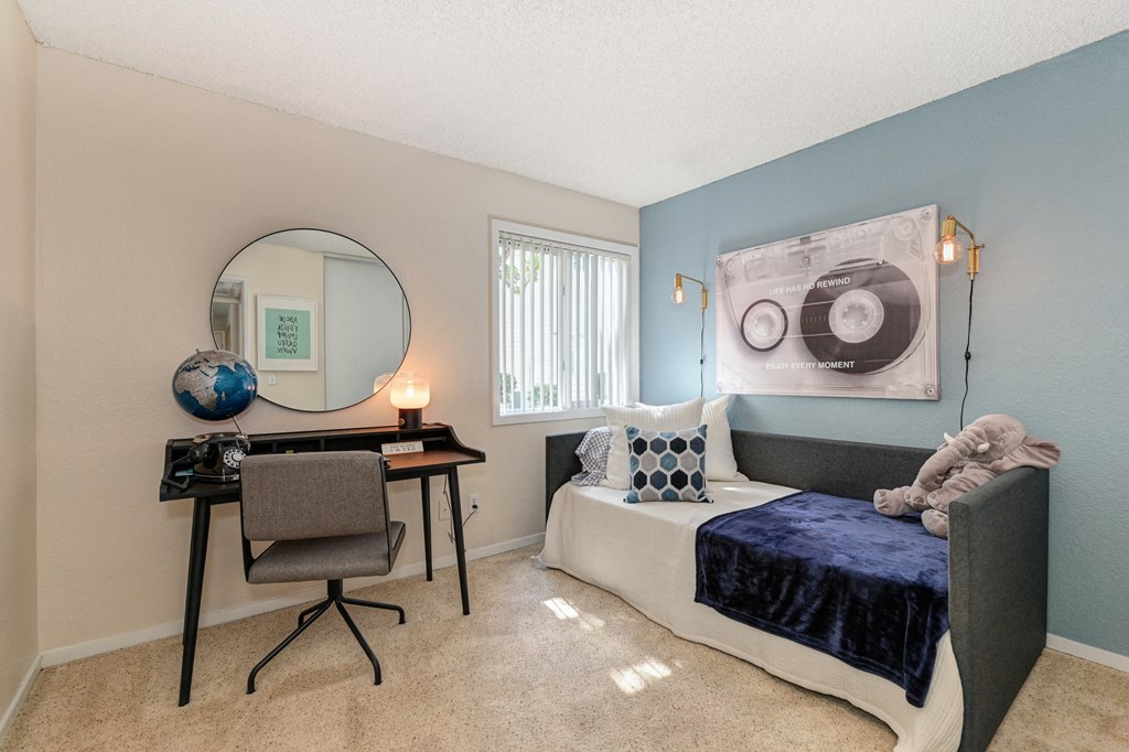 a bedroom with a daybed along the wall and a desk with mirror on the wall.  Bedroom also has carpet and window.