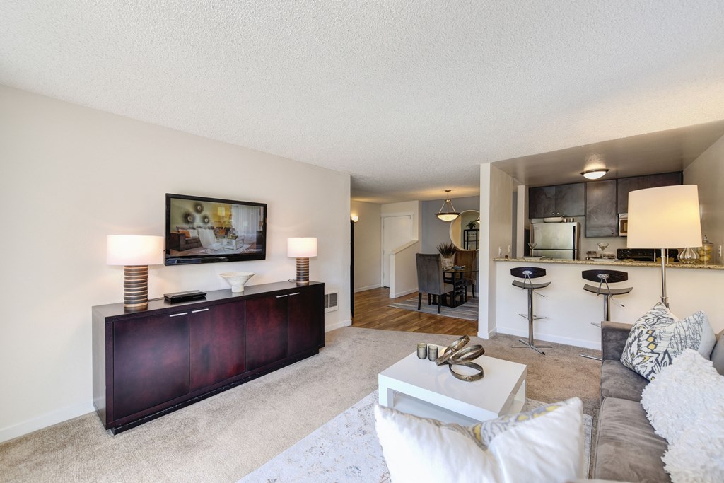 Model Living room with gray couch and white chair.  Kitchen and counter with bar stools is visible from the living room at The Retreat at Walnut Creek, California, 94596