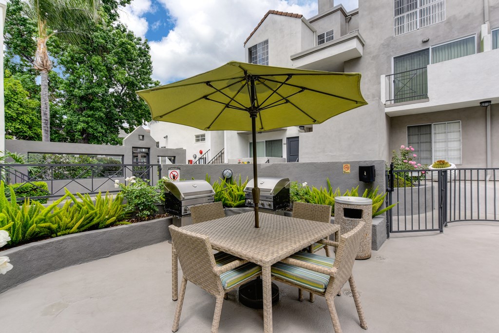 Outdoor table with chairs and lime green shade umbrella. Green shrubs in planter around the seating area and gas BBQ's.