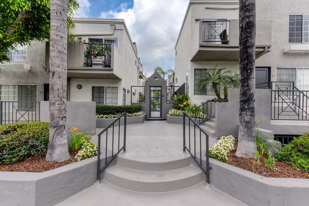 Apartment complex entry from the street with 2 stairs leading up to the community entry door. Palm trees are on both sides of the stairs. 