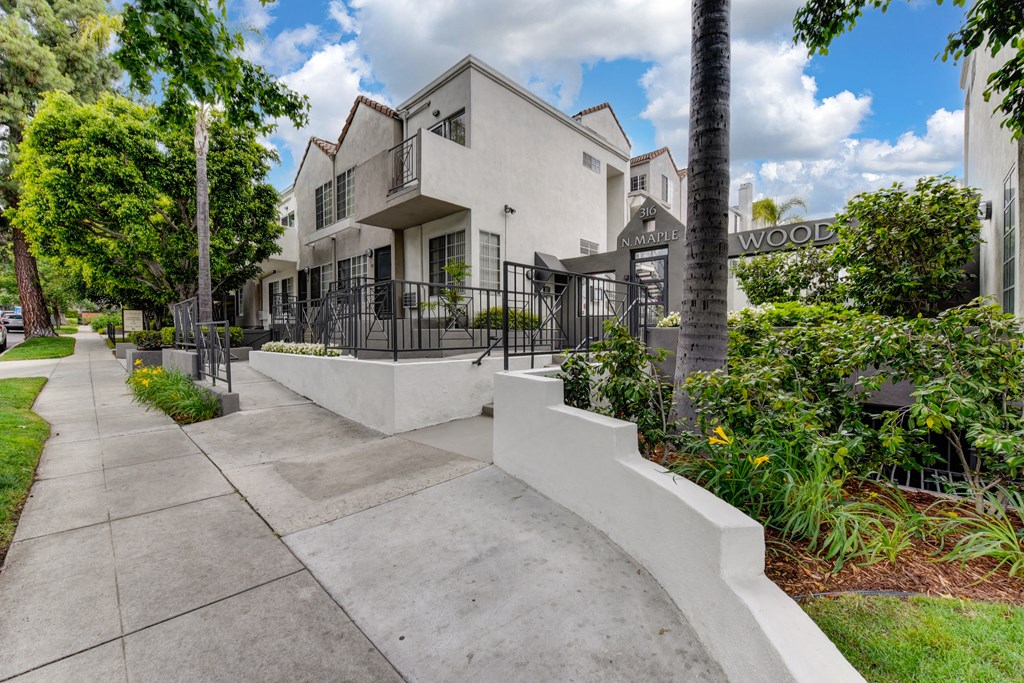Street sidewalk leading up to the community entrance.  Green pants and trees outside of the entrance. 