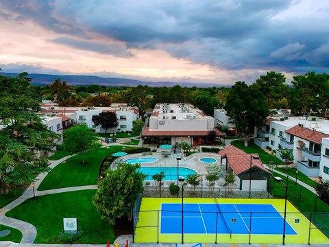 A tennis court is surrounded by a pool and apartment buildings.
