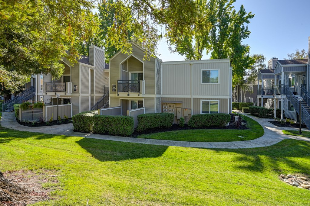 Exterior of the apartment community with large grassy area, mature trees and landscaped bushes at Rush River Apartments, California.