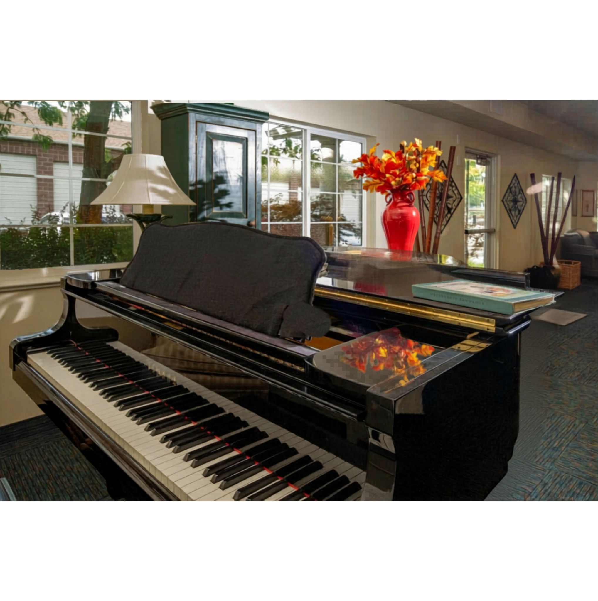 A black grand piano with a black cover sits in a room with a red vase of flowers on top of it.