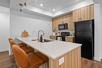 A kitchen with a black refrigerator, brown chairs, and wooden cabinets.