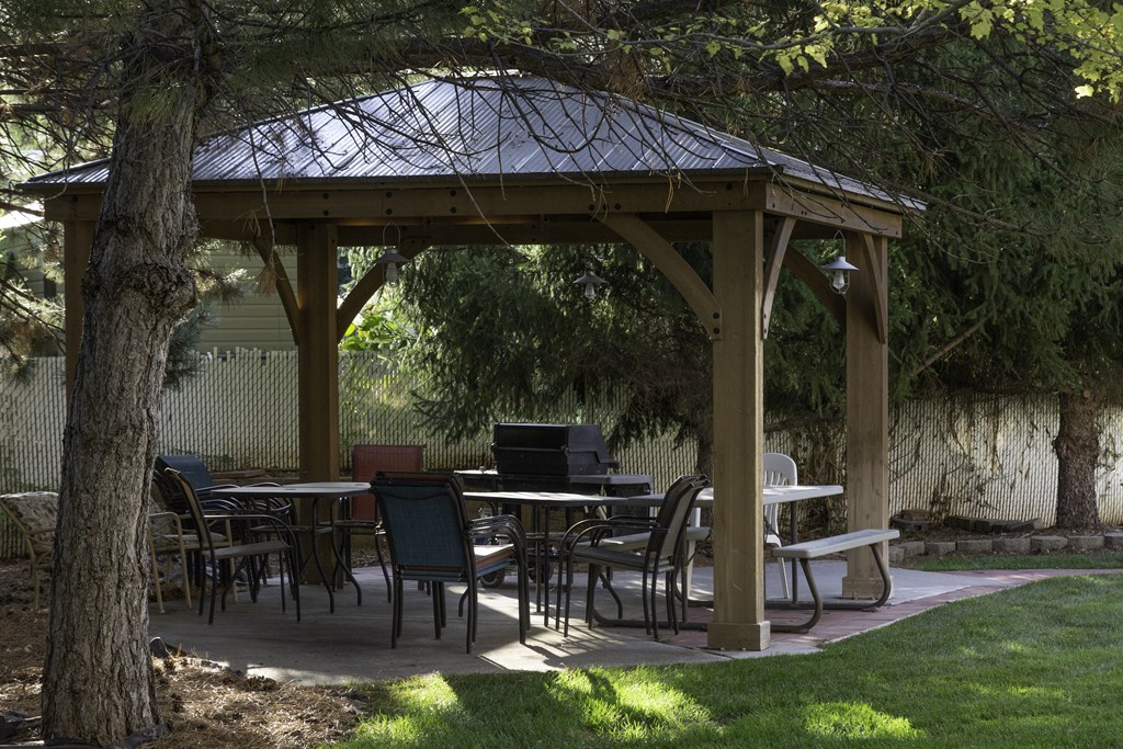 A wooden gazebo with a metal roof is surrounded by trees and has tables and chairs set up for outdoor dining.