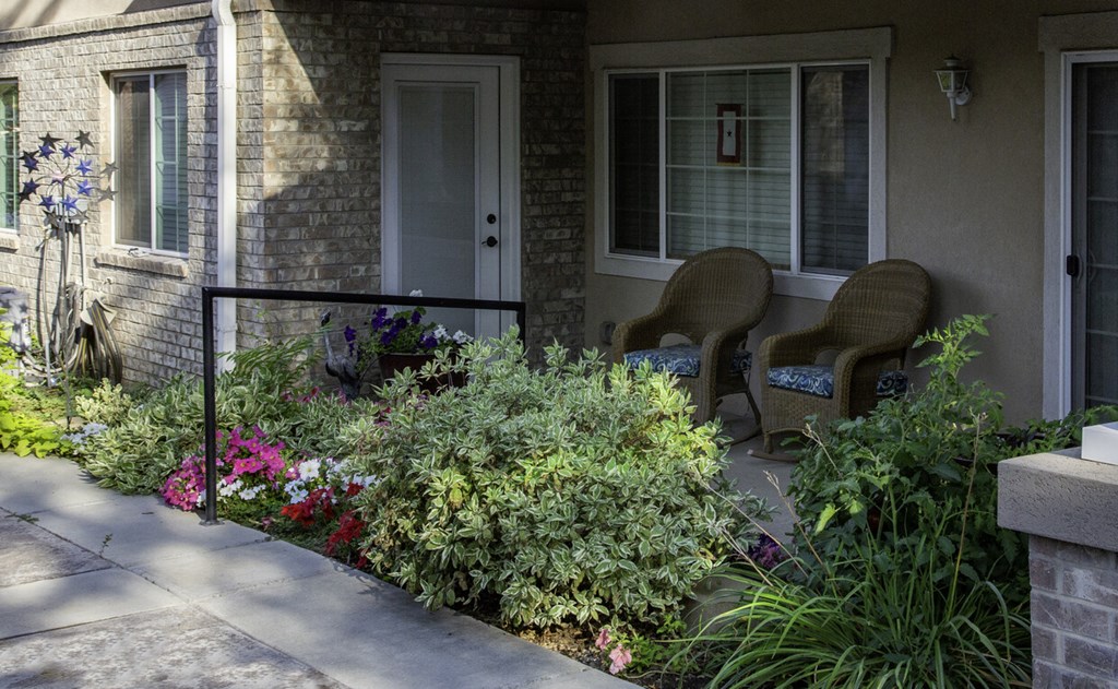 Two chairs are on a porch with a flower bed in front.