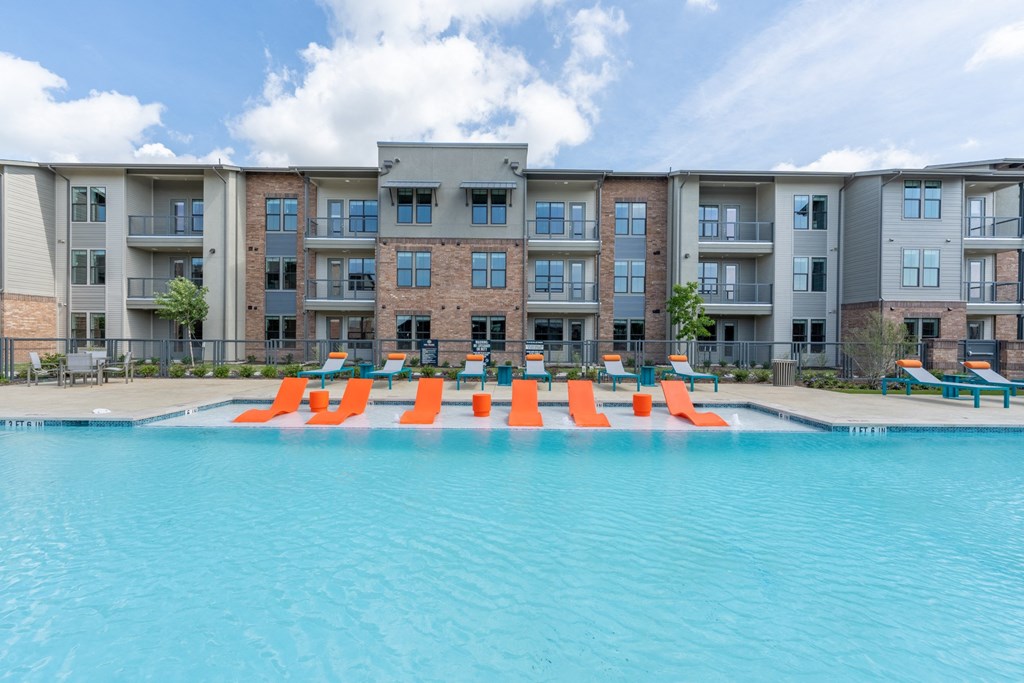a swimming pool with orange lounge chairs in front of an apartment building at One Preston Station, Celina, TX, 75009
