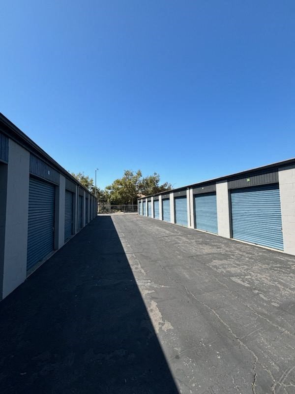 Outdoor storage facility with multiple units featuring blue roll-up doors and an asphalt driveway under a clear blue sky.