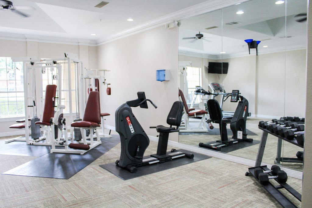 Modern-style fitness center with huge mirror, tan flooring, free weights and cardio machines at Montfort Place Apartments, Texas