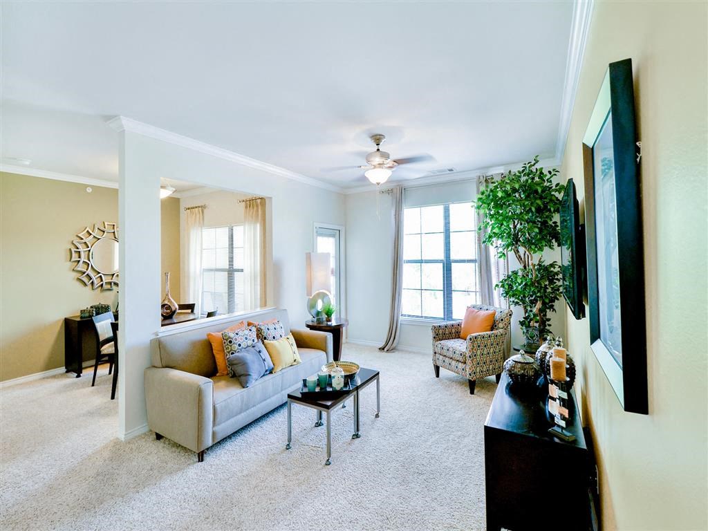 Large living room with cream carpeting, a beige couch, a tan chair and tall windows that provide natural light at Mission at La Villita Apartments, Irving, TX