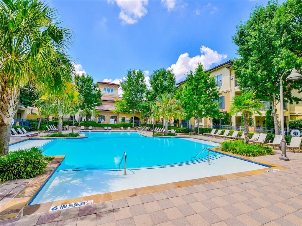 Large swimming pool surrounded by trees, tan stone, and white lounge chairs at Mission at La Villita Apartments, Irving 75039