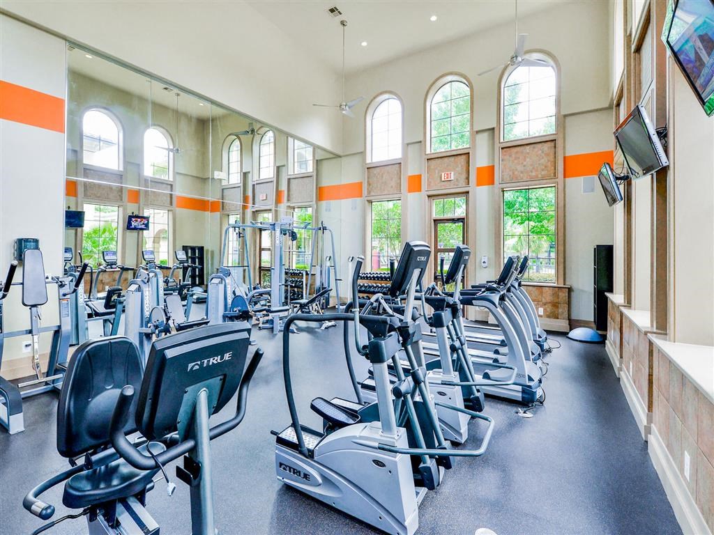 Fully equipped fitness center with cardio equipment, soaring windows, black flooring and tan walls at Mission at La Villita Apartments, Texas