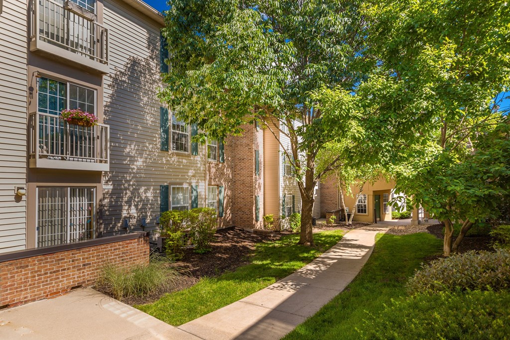 A tree-lined walkway leads to a building with a brick pillar at Prairie Commons - 55+ Senior Community Apartments, Lawrence