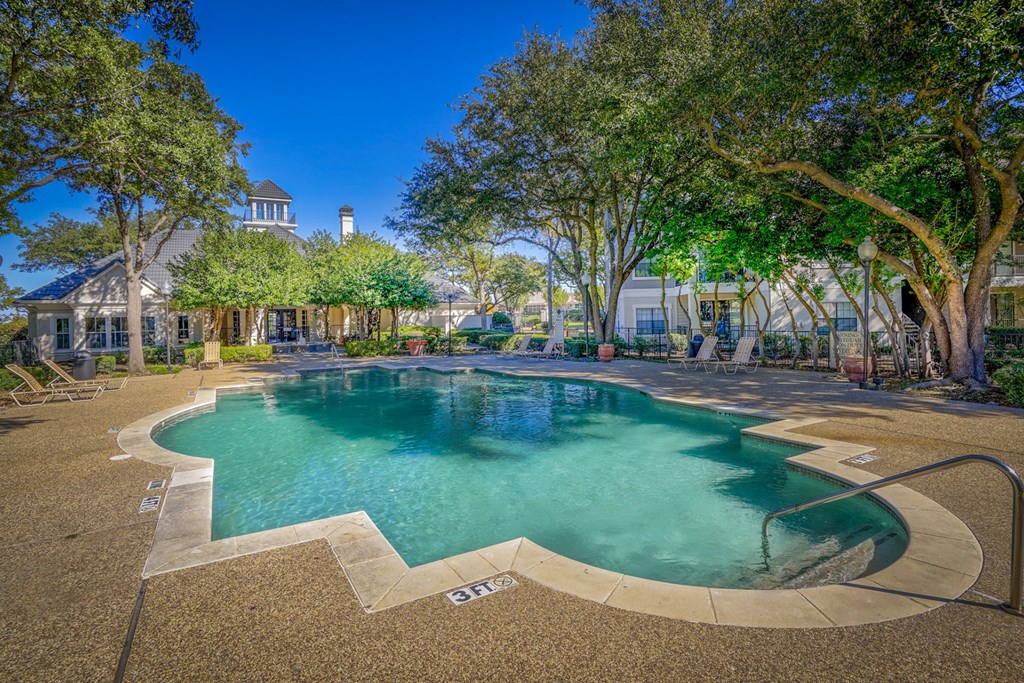 Outdoor pool area at Saxony at Chase Oaks Apartments in Dallas, TX, featuring a clear turquoise swimming pool, a sun deck with lounge chairs, mature trees for shade, and a charming clubhouse in the background with a turret-style roof.