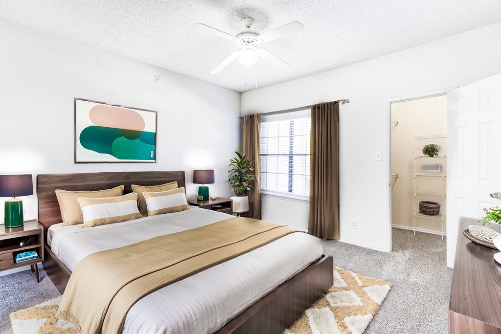 A staged bedroom at Saxony at Chase Oaks Apartments in Dallas, TX, showcasing a modern bed with stylish bedding, side tables, a ceiling fan, and a walk-in closet with shelving.