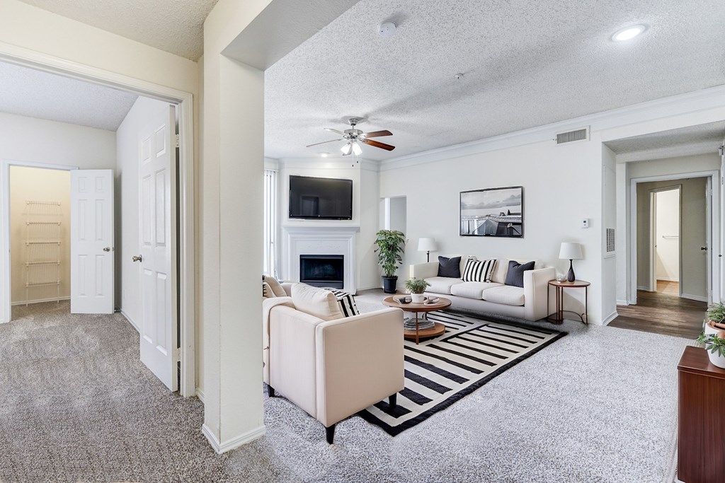 Spacious living room in Saxony at Chase Oaks Apartments in Dallas, TX, featuring a staged modern design with neutral-colored sofas, striped area rug, fireplace with a mounted TV, and large windows for natural light.