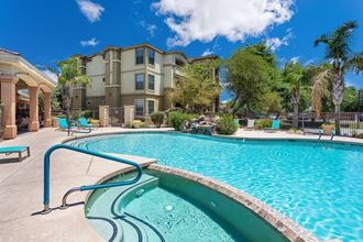 Photo of a large swimming pool and spa. Blue pool chairs line the sides of the pool 3 story apartment building is seen in the background