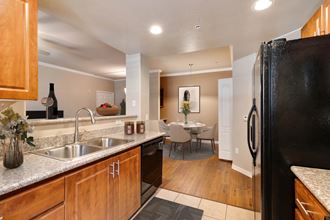Photo of kitchen looking out to dining area with roud table and four charis. Stainless steel double sink and gloss black dishwasher can be seen on the left with a gloss black double door refrigerator to the right