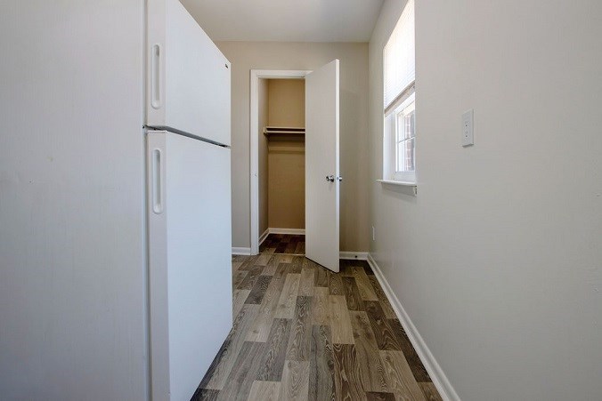 A white refrigerator is in a narrow hallway with wood flooring.