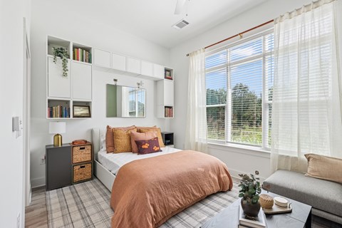 A bedroom with a bed, a window with blinds, a nightstand, a bookshelf, and a plant on a table at Link Apartments® CYKEL Apartments, Charlotte, NC, 28208