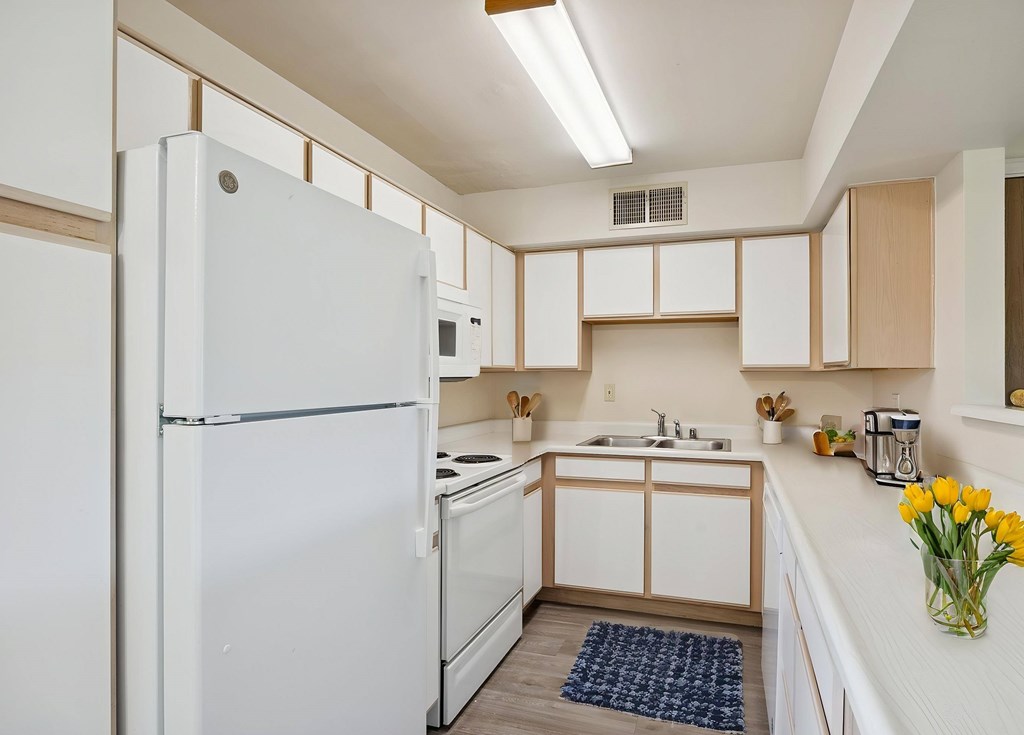 A white refrigerator stands in a kitchen with wooden cabinets and a sink.