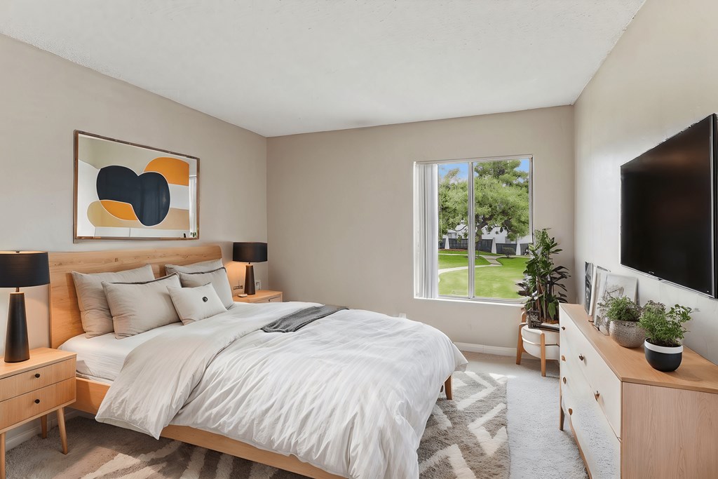 A bedroom with a large bed, a nightstand, and a window overlooking a green lawn.