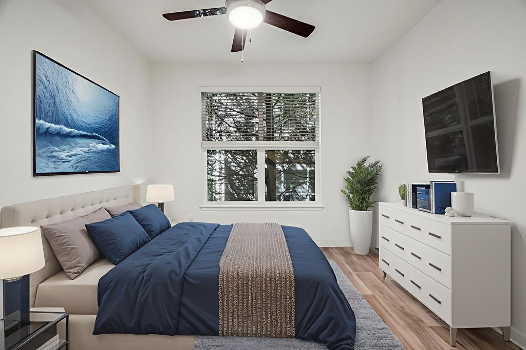 A bedroom with a bed, a ceiling fan, a window with a view of trees, a nightstand with a lamp, and a painting on the wall.
