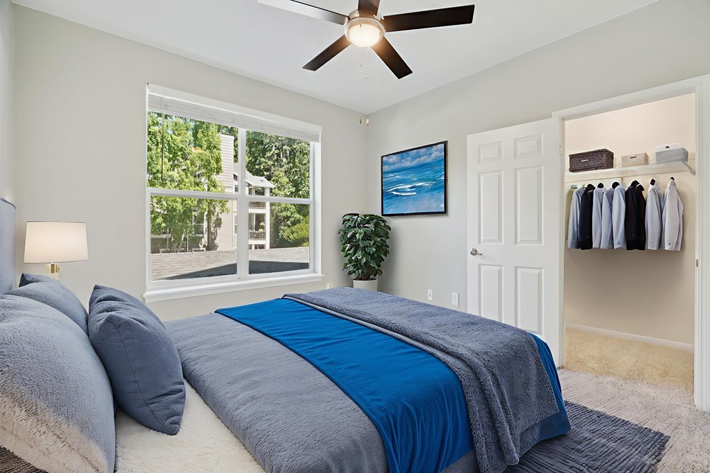 A bedroom with a bed, a ceiling fan, and a window with a view of a tree-filled outdoor area.