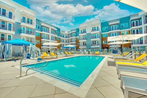 A swimming pool surrounded by sun loungers and umbrellas in front of a multi-story hotel.