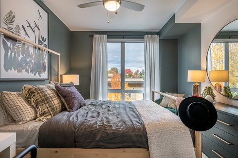 A bedroom with a bed, a hat on the bed, a painting on the wall, a window with a view of trees, and a lamp on the side table.