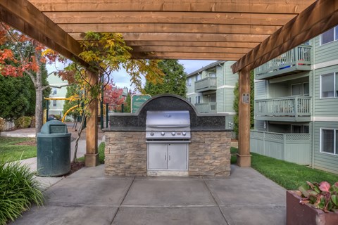 A stone and tile outdoor kitchen with a grill and sink under a wooden pergola.