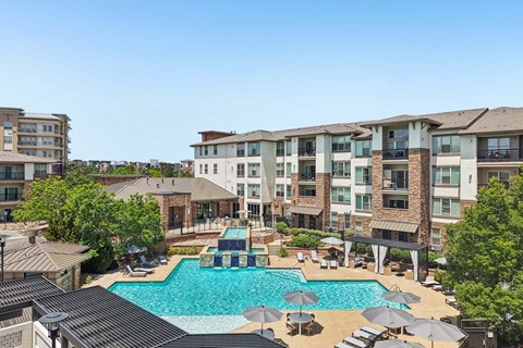A large swimming pool surrounded by lounge chairs and umbrellas in front of a multi-story apartment building.