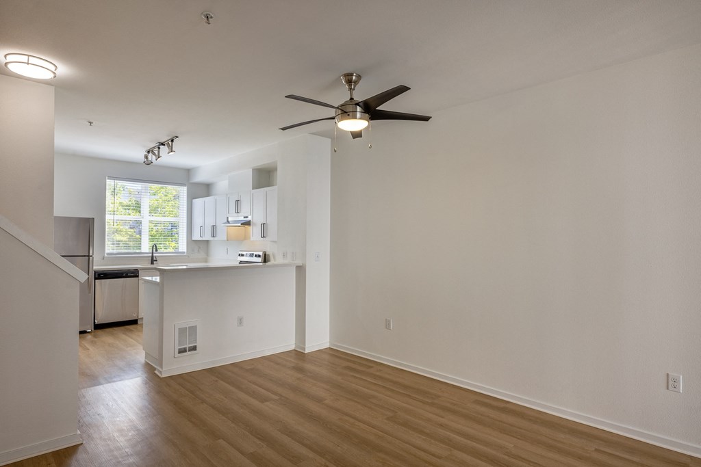 an empty living room with a ceiling fan and a kitchen in the background