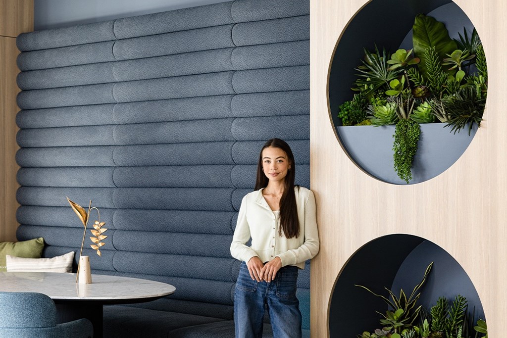 A woman in a white shirt and blue jeans stands in front of a grey wall with a round mirror and a potted plant.