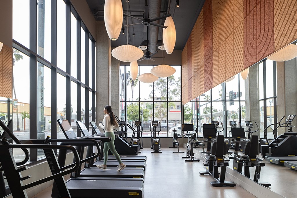 A woman is running on a treadmill in a gym with large windows.