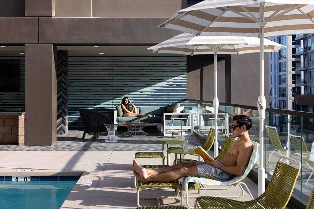 A man is sitting on a chair reading a book under an umbrella on a patio.