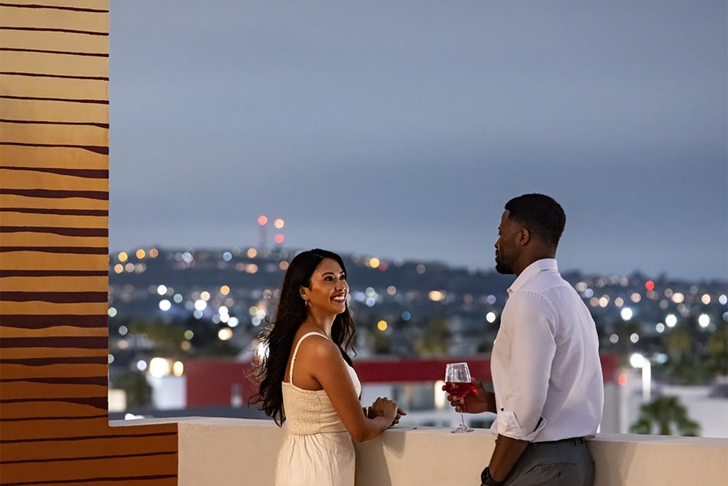 A couple standing on a balcony with a cityscape view at dusk.