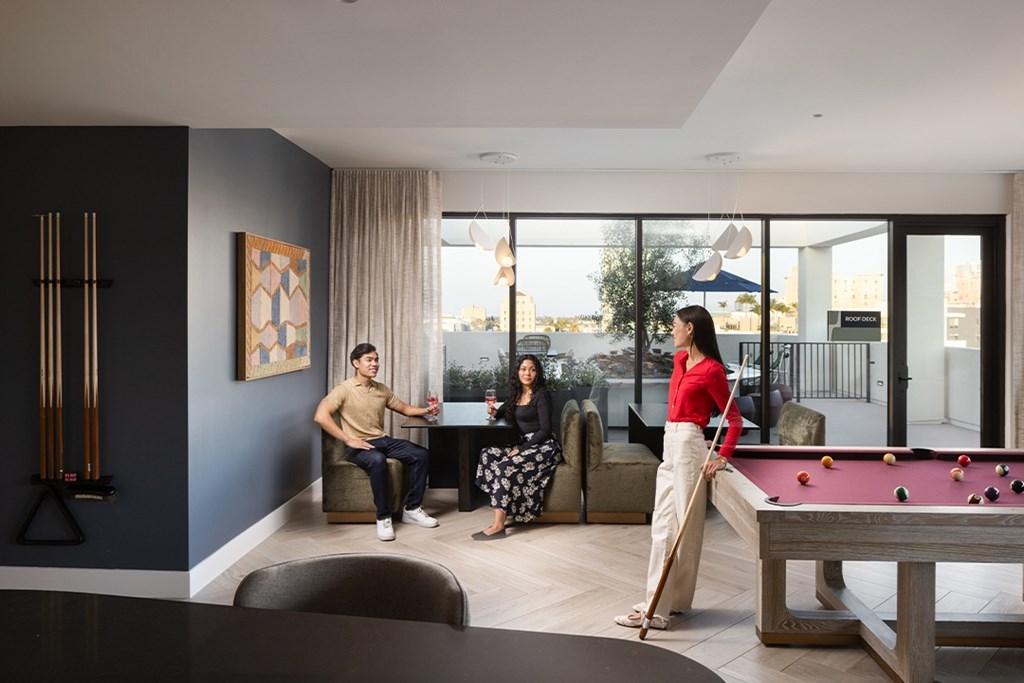 Three people are sitting and standing around a pool table in a room.