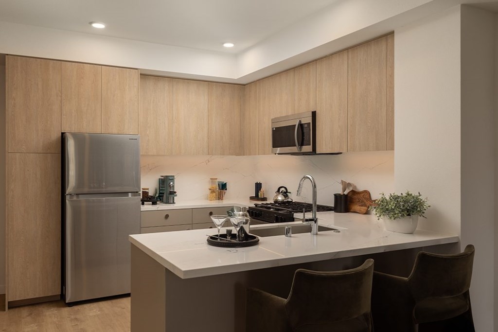 A modern kitchen with a stainless steel refrigerator, a white countertop, and brown chairs.