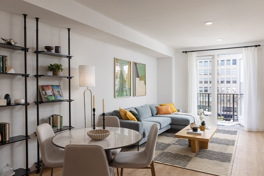 A modern living room with a glass table and chairs, a grey sofa, and a view of a balcony.