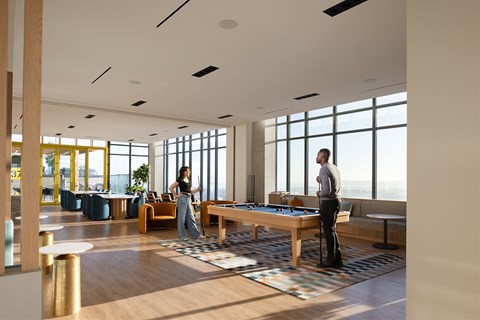 Two people playing pool in a brightly lit room with large windows.
