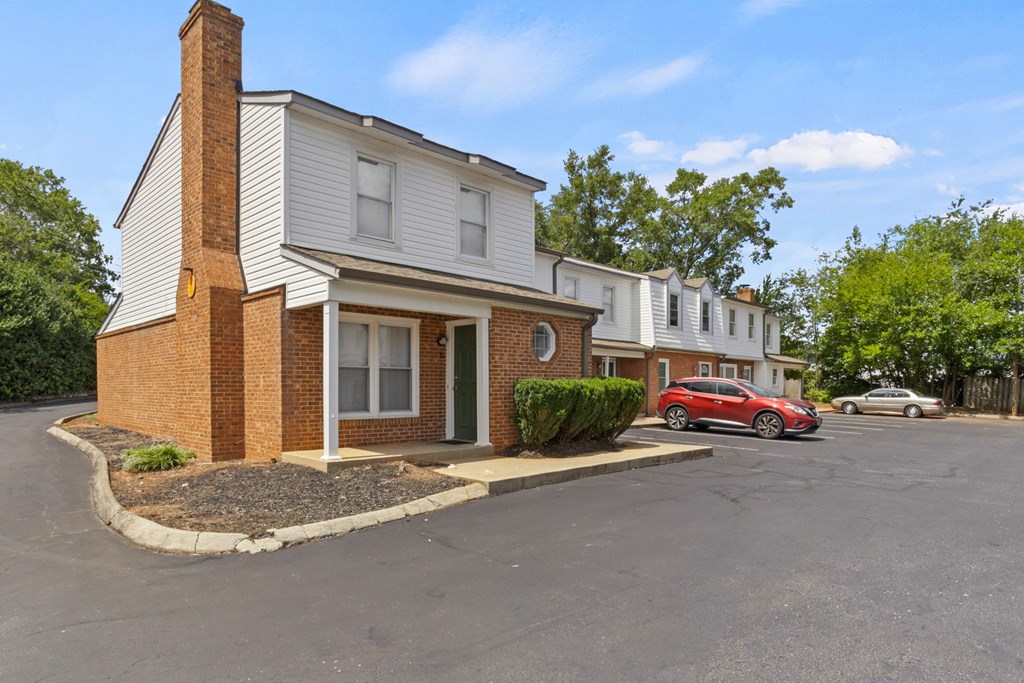 A red car is parked in front of a white house with a brick chimney.
