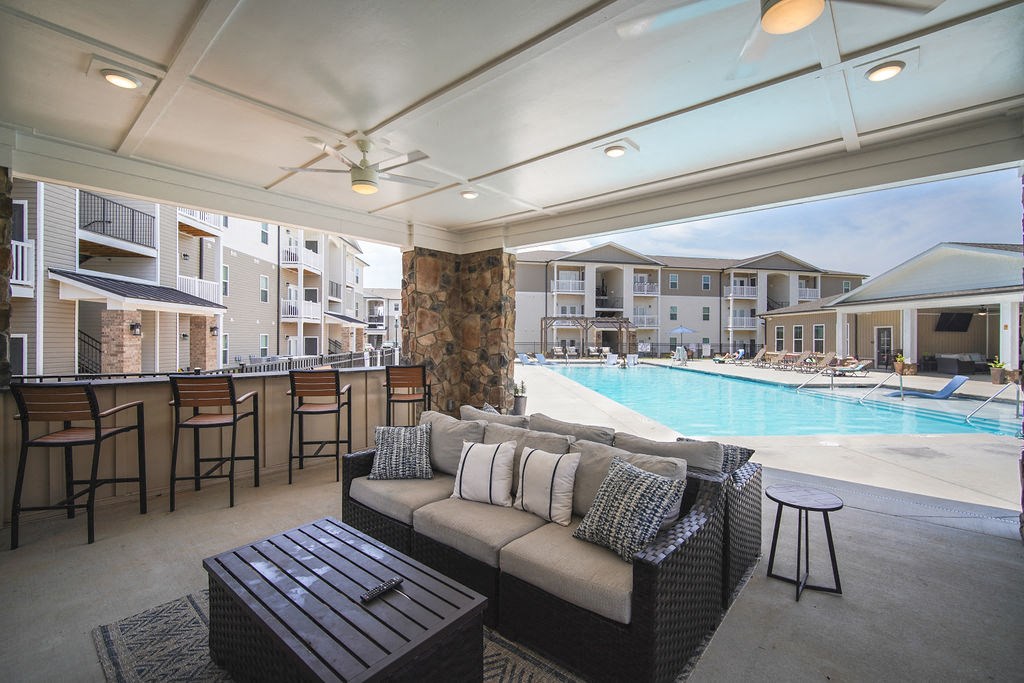 Pet-Friendly Apartments in Boiling Springs, SC - Peachtree Village - Outdoor Patio with Two Rattan Couches, Coffee Table, Stool, Bar Chairs, Ceiling Fans, and a View of the Swimming Pool Area.