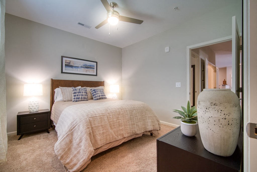 One Bedroom Apartments in Boiling Springs, SC - Peachtree Village - Bedroom with Carpet Flooring, Bed, Nightstand, Lamps, Ceiling Fan, Wall Frame, and Vase.