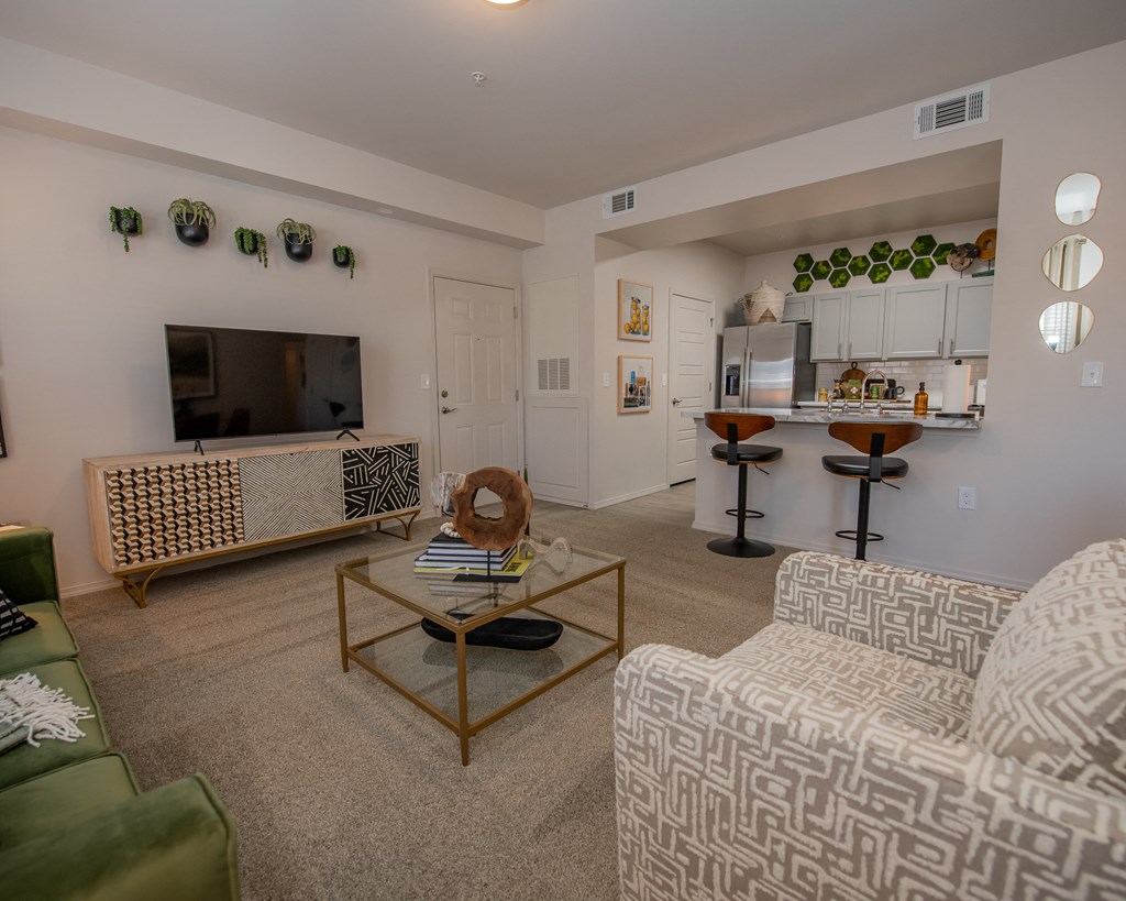 A living room with a television, a sofa, a coffee table, and a kitchen area in the background.